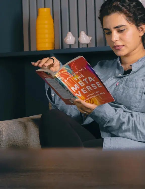 An Asian girl wearing a blue shirt and black jeans; sitting and reading ‘The Meta-verse’ with 2 porcelain birds and a yellow-mustard vase on a blue shelve in the background.